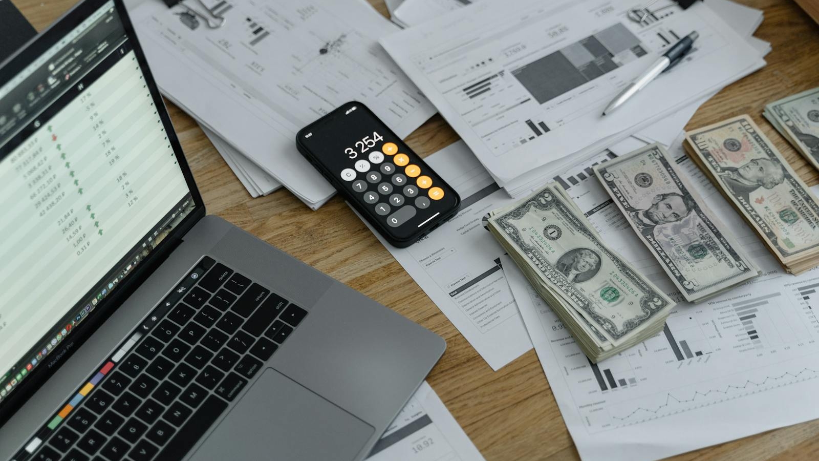 A desk with financial documents, currency, a laptop, and phone calculator.